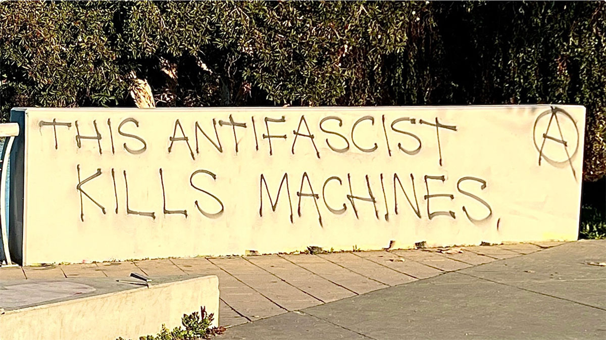 The image shows a concrete wall with graffiti written on it. The graffiti reads “THIS ANTIFASCIST KILLS MACHINES” in capital letters.